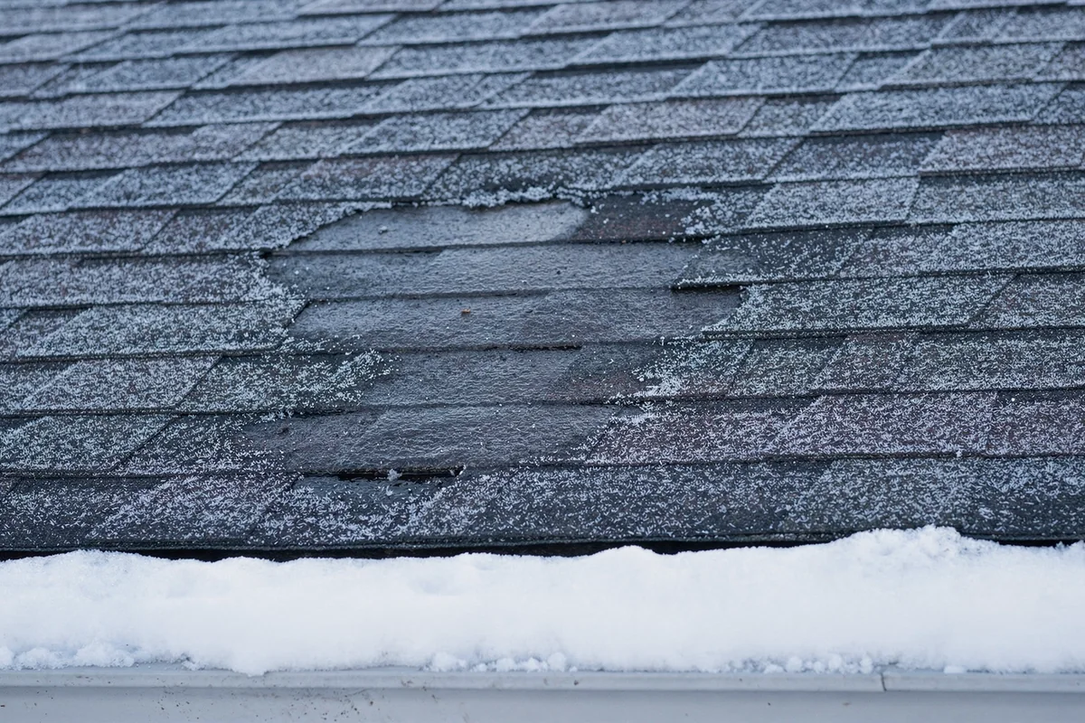 A frosted shingle slope with one bare melted patch, above an overhang still heaped with snow.